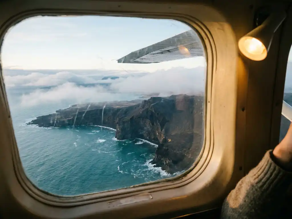 Uitzicht vanuit vliegtuigraam op IJslands vulkanisch kustlandschap met zwarte kliffen en turquoise Atlantische oceaan.