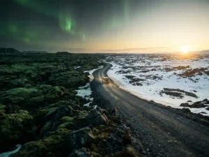 Smal grindweggetje door IJslands lavaveld met mos en sneeuw, noorderlicht in groene tinten boven gouden middernachtzon aan de horizon.