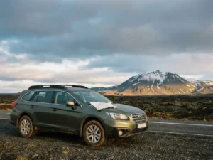 Huurauto geparkeerd langs IJslands Ring Road met zwarte lavavelden, besneeuwde vulkaan en wegenkaart op de motorkap tijdens gouden uur.