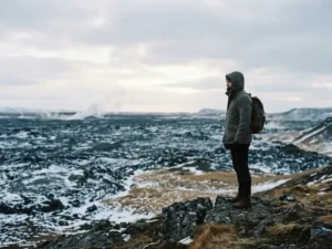 Reiziger aan de rand van IJslands lavaveld in het laagseizoen, met besneeuwd zwart gesteente onder een bewolkte winterlucht.