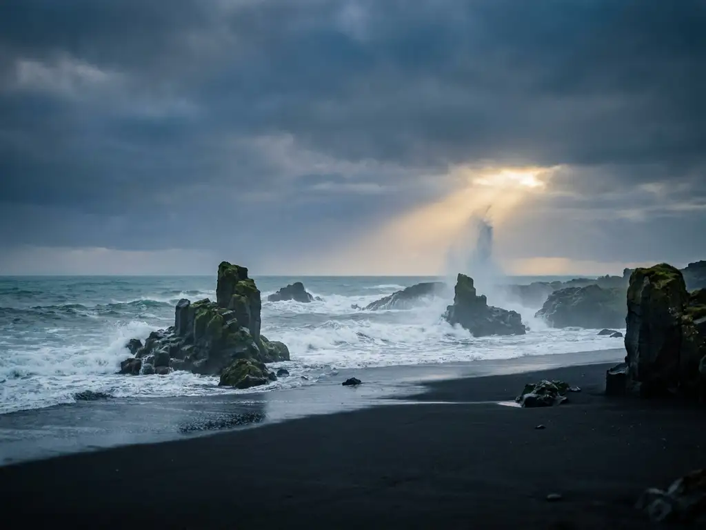 Zwart vulkanisch zandstrand in IJsland met basaltrotsen en schuimende Atlantische oceaan onder bewolkte lucht met gouden lichtstraal.