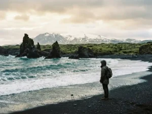 Reiziger op zwart lavastrand in IJsland met basaltrotsen in de branding en besneeuwde vulkanische bergrug op de achtergrond.
