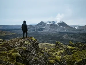 Toerist staat aan de rand van een IJslands vulkaangebied, met zwarte lavavelden en een stomende krater op de achtergrond.