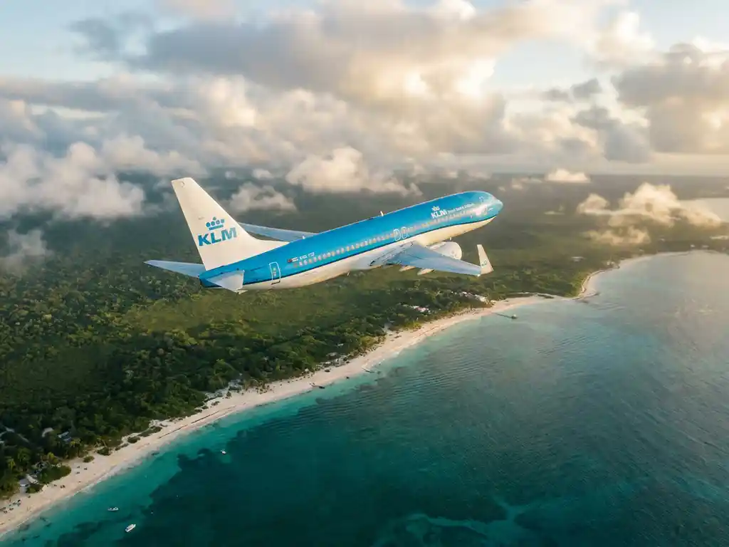 KLM-vliegtuig boven turquoise Caribische kustlijn met wit zandstrand, weelderige jungle en gouden zonlicht door wolken.