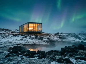 Luxe glazen lodge op met sneeuw bedekte IJslandse heuvel, met gouden binnenverlichting, dampende geothermische bron en noorderlicht aan schemerende hemel.