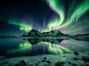 Groen en violet noorderlicht boven IJslands gletsjermeer, weerspiegeld in het stille water met besneeuwde vulkanen aan de horizon.