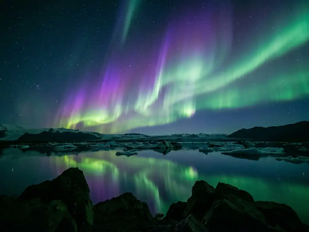 Groene en violette noorderlichten boven IJslands gletsjermeer, weerspiegeld in het stille water met zwarte vulkanische rotsen op de voorgrond.