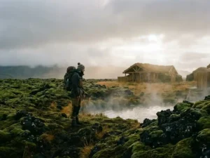 Reiziger naast met mos bedekt lavaveld in IJsland, geothermische stoomwolken en houten eco-lodge zichtbaar op de achtergrond.