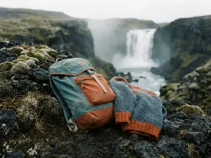 Verweerde wandelrugzak en grove wollen trui op zwart vulkanisch gesteente, omringd door Arctisch mos met IJslandse waterval op de achtergrond.