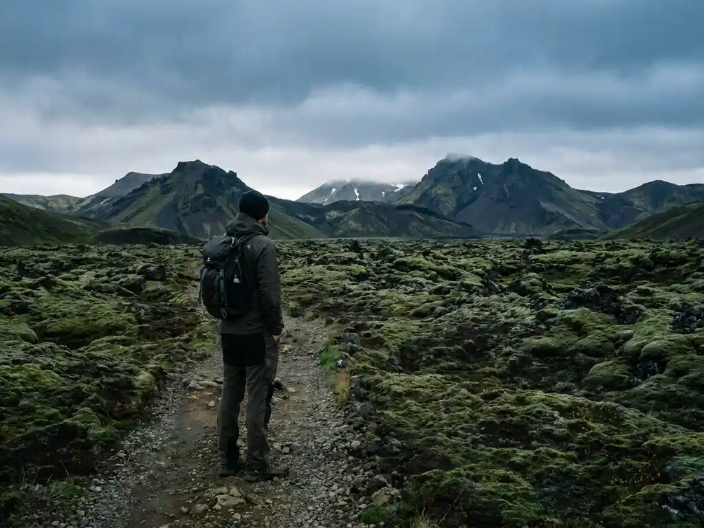 Wandelaar op smal grindpad langs IJslands mosbegroeid lavaveld, met vulkanische bergen en grijze lucht op de achtergrond.