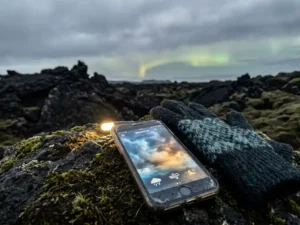 Weerapp op verweerde smartphone naast wollen handschoen op vulkanisch lavagesteente, IJslands stormlandschap met hint van noorderlicht.