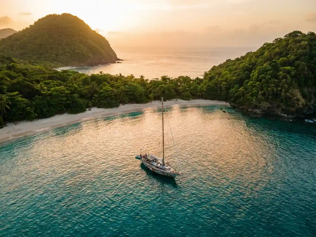 Houten zeilboot voor anker in turquoise Caribische baai tussen vulkanische kustlijnen met wit zandstrand en tropisch groen.