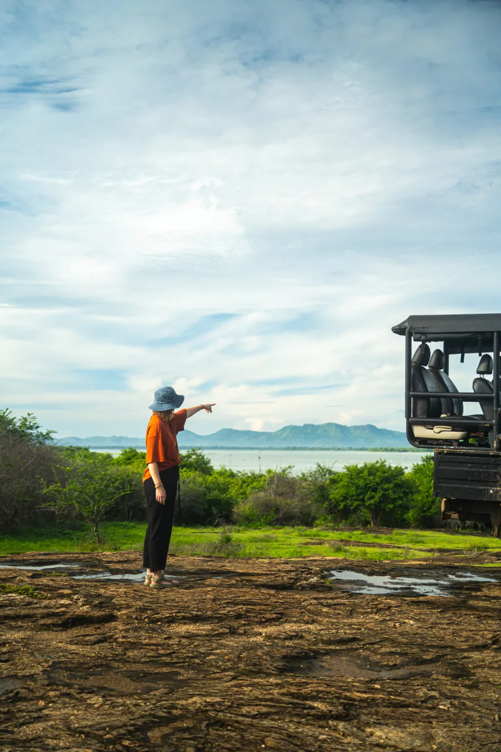Een reiziger met een hoed en oranje shirt staat naast een safari-jeep op een open veld en wijst naar de verte onder een lichtbewolkte lucht in Udawalawe.