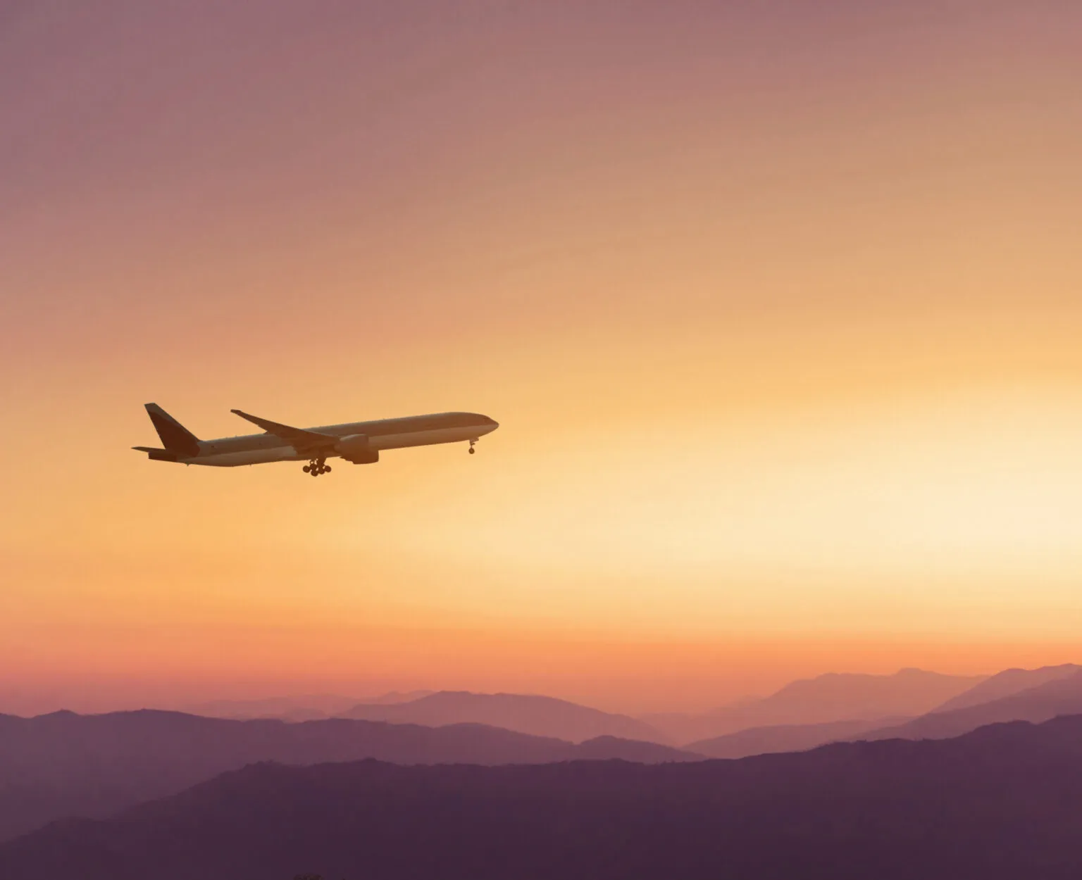 Silhouet van een opstijgend passagiersvliegtuig in een oranje-roze lucht boven bergen tijdens zonsondergang.