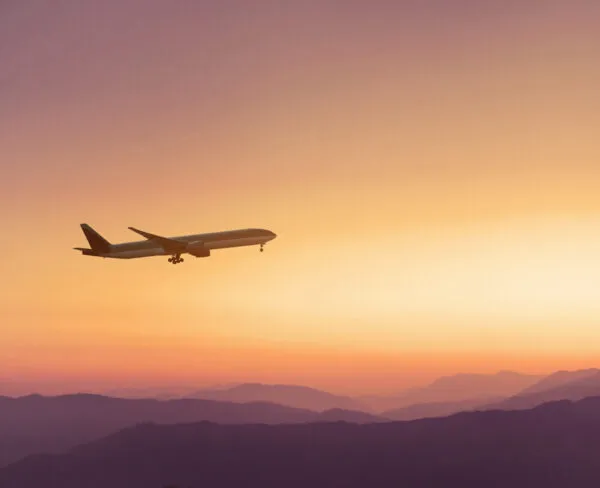 Silhouet van een opstijgend passagiersvliegtuig in een oranje-roze lucht boven bergen tijdens zonsondergang.
