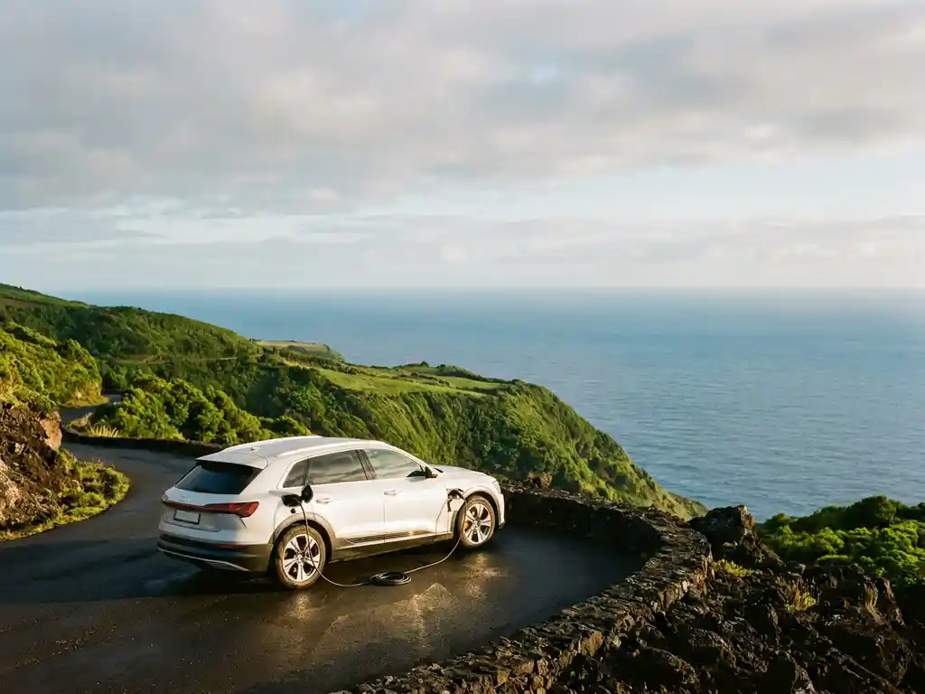 Witte elektrische auto met laadkabel op vulkanische kustweg op de Azoren, omgeven door groene heuvels en de Atlantische Oceaan.