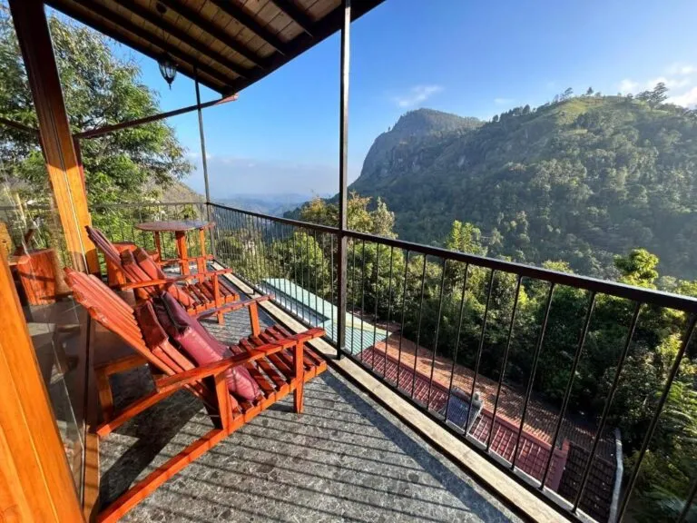 Twee rode houten ligstoelen staan op een overdekt balkon met een zwarte leuning, uitkijkend op groene, bergachtige heuvels onder een helderblauwe lucht in Ella.